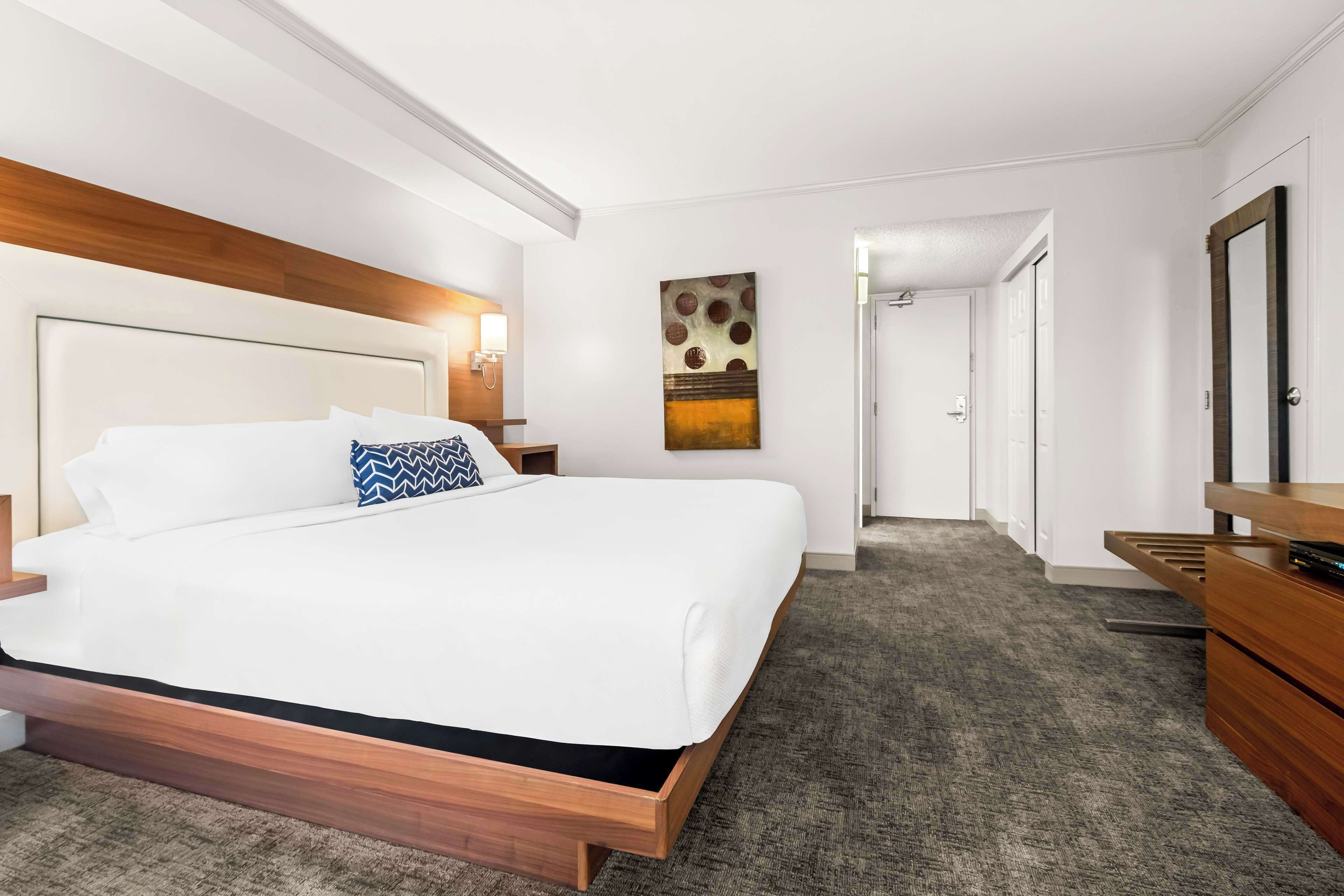 A modern hotel room with a large bed, wooden headboard, a nightstand, a dresser, and a painting on the wall.