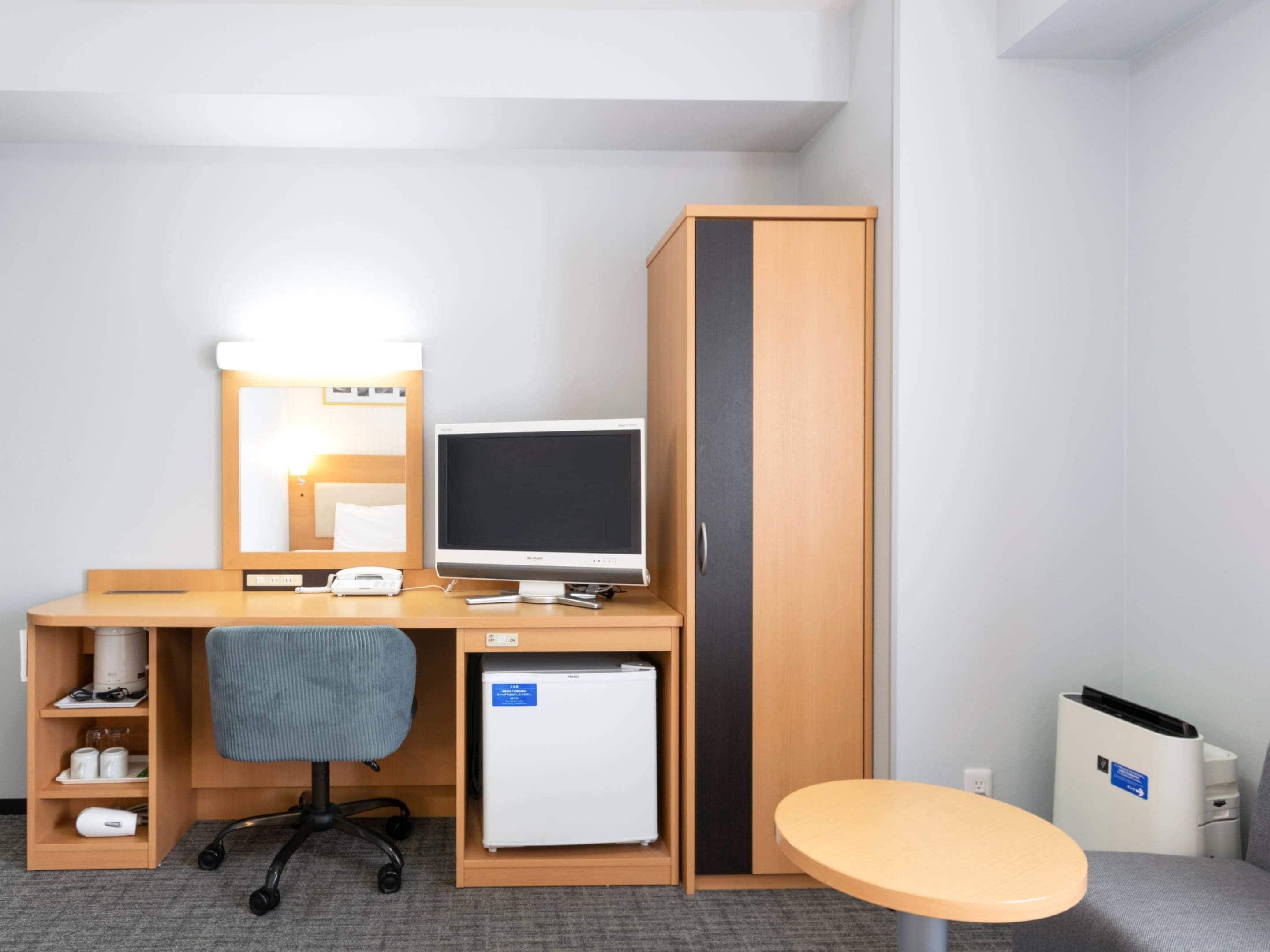Habitación de hotel pequeña con escritorio de madera, refrigerador, lámpara de escritorio, silla y mesita.