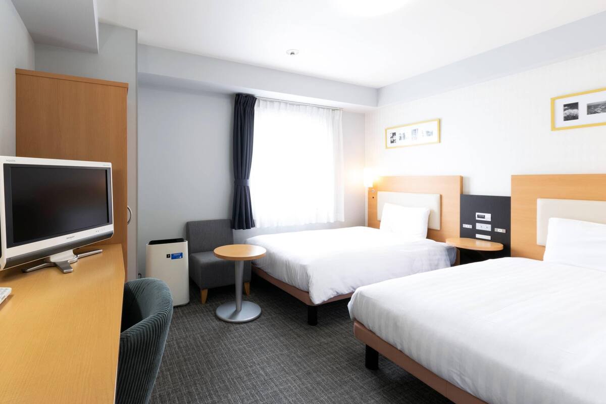 Ein Hotelzimmer mit zwei Betten, einem Schreibtisch, einem Flachbildfernseher, einem kleinen runden Tisch und einem grauen Sessel.