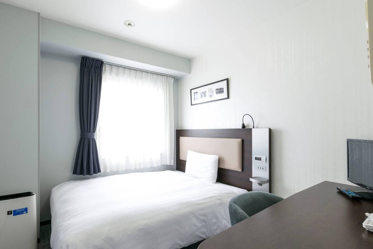 Ein Hotelzimmer mit Bett, Schreibtisch, Stuhl, einem Fenster mit Vorhängen, einem an der Wand befestigten Fernseher und einer Bedienkonsole.