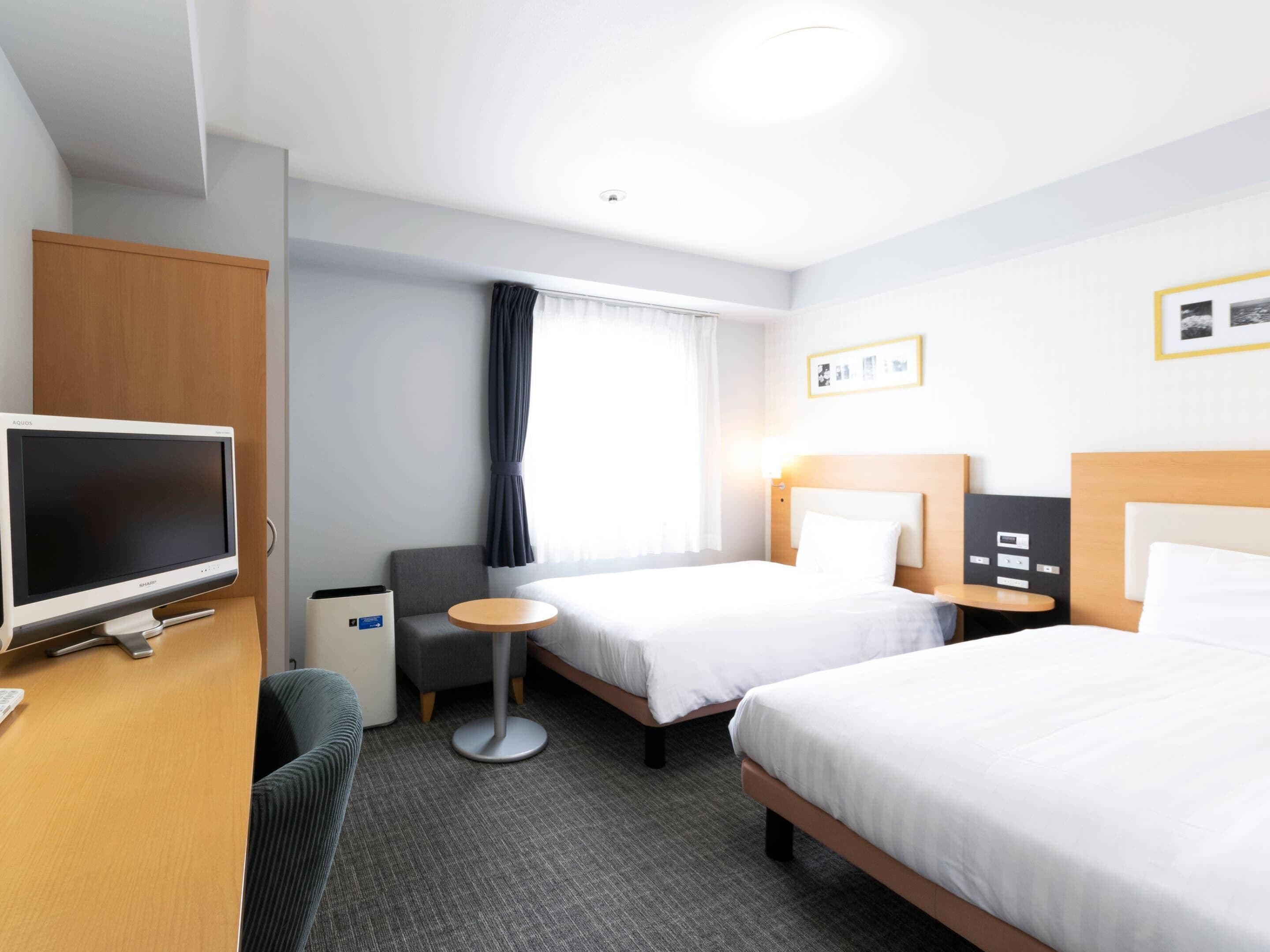 A hotel room with two beds, a desk with a flat-screen TV, a small round table, and a grey armchair.