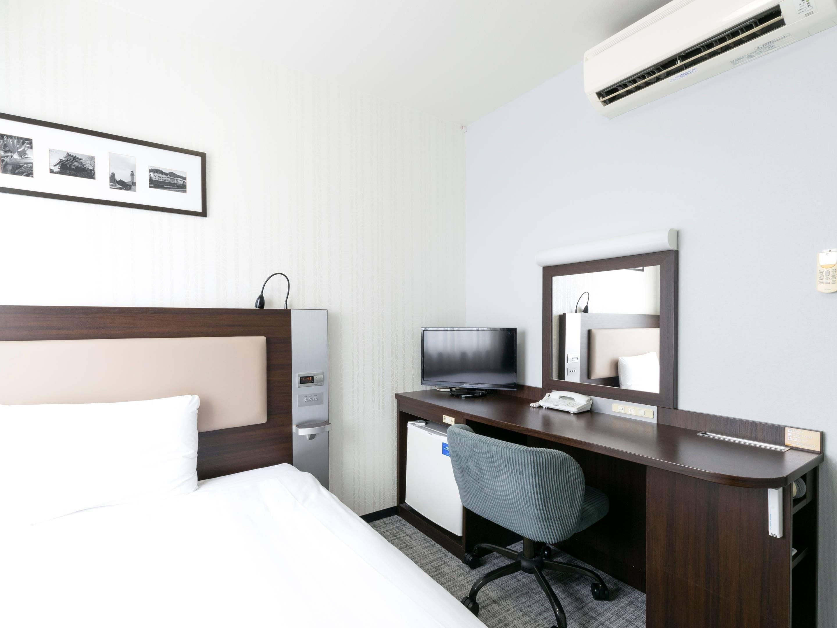 A hotel room with a bed, a desk with a chair, a television, a mirror, and an air conditioning unit.
