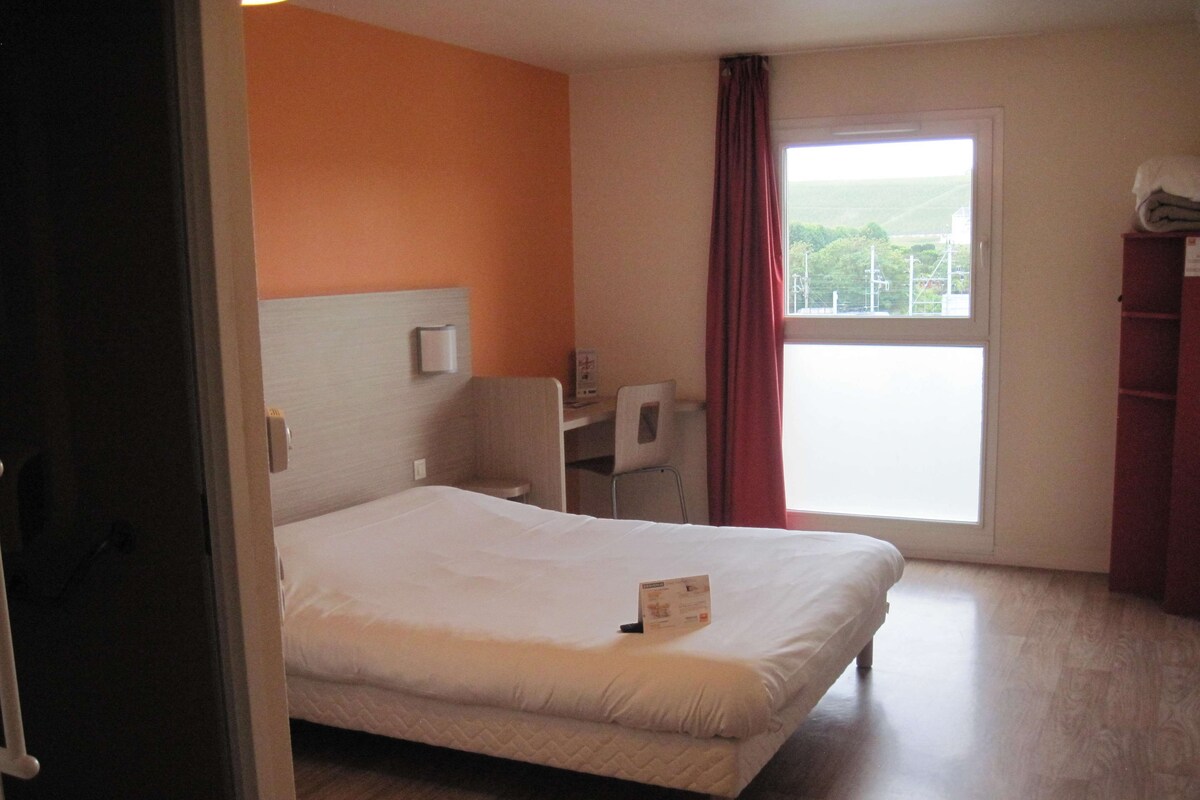 A hotel room with a bed, a desk, and a window with a view of the countryside.