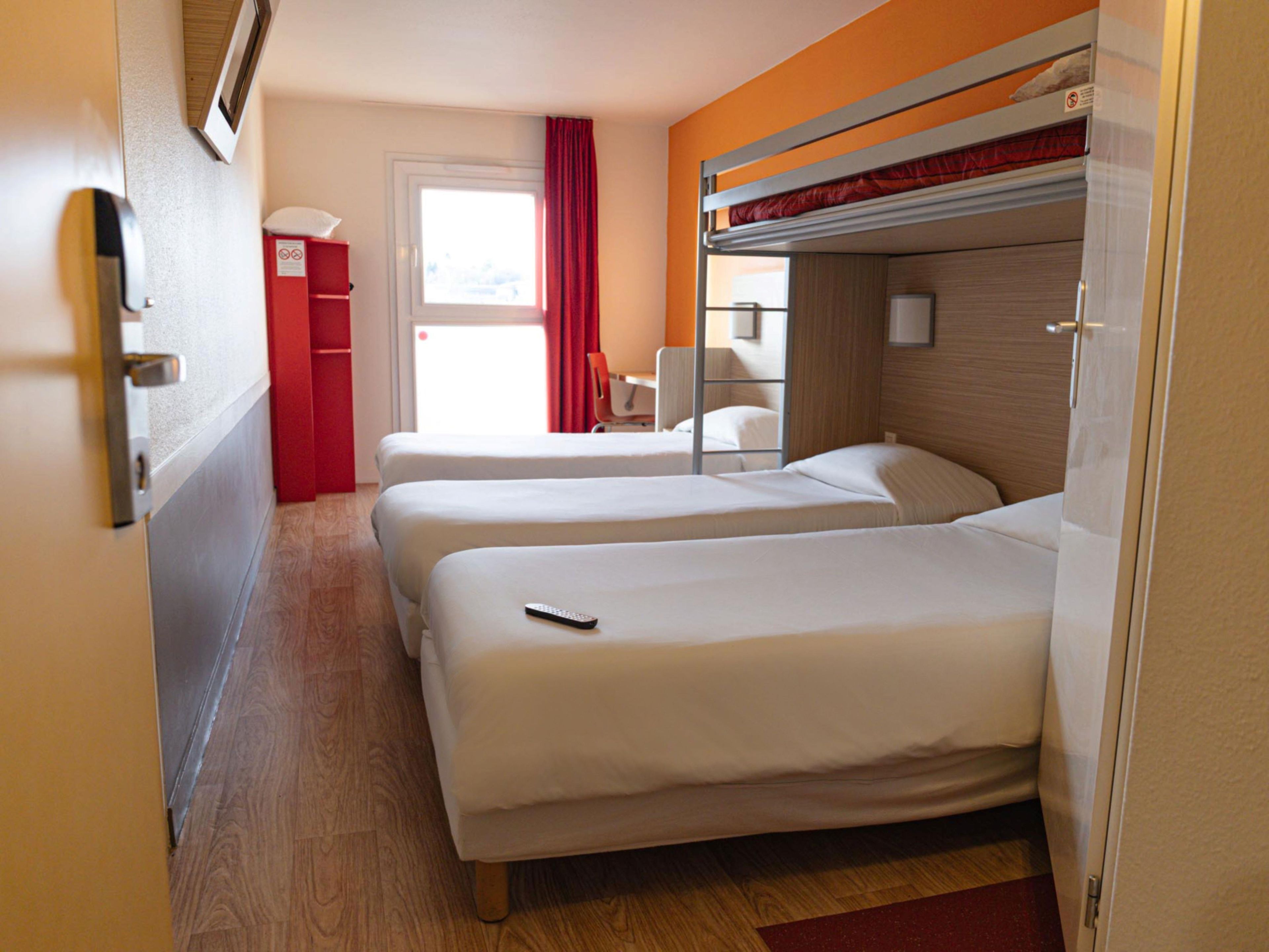 A hotel room with two beds, a red cabinet, a window with curtains, and a bunk bed above.