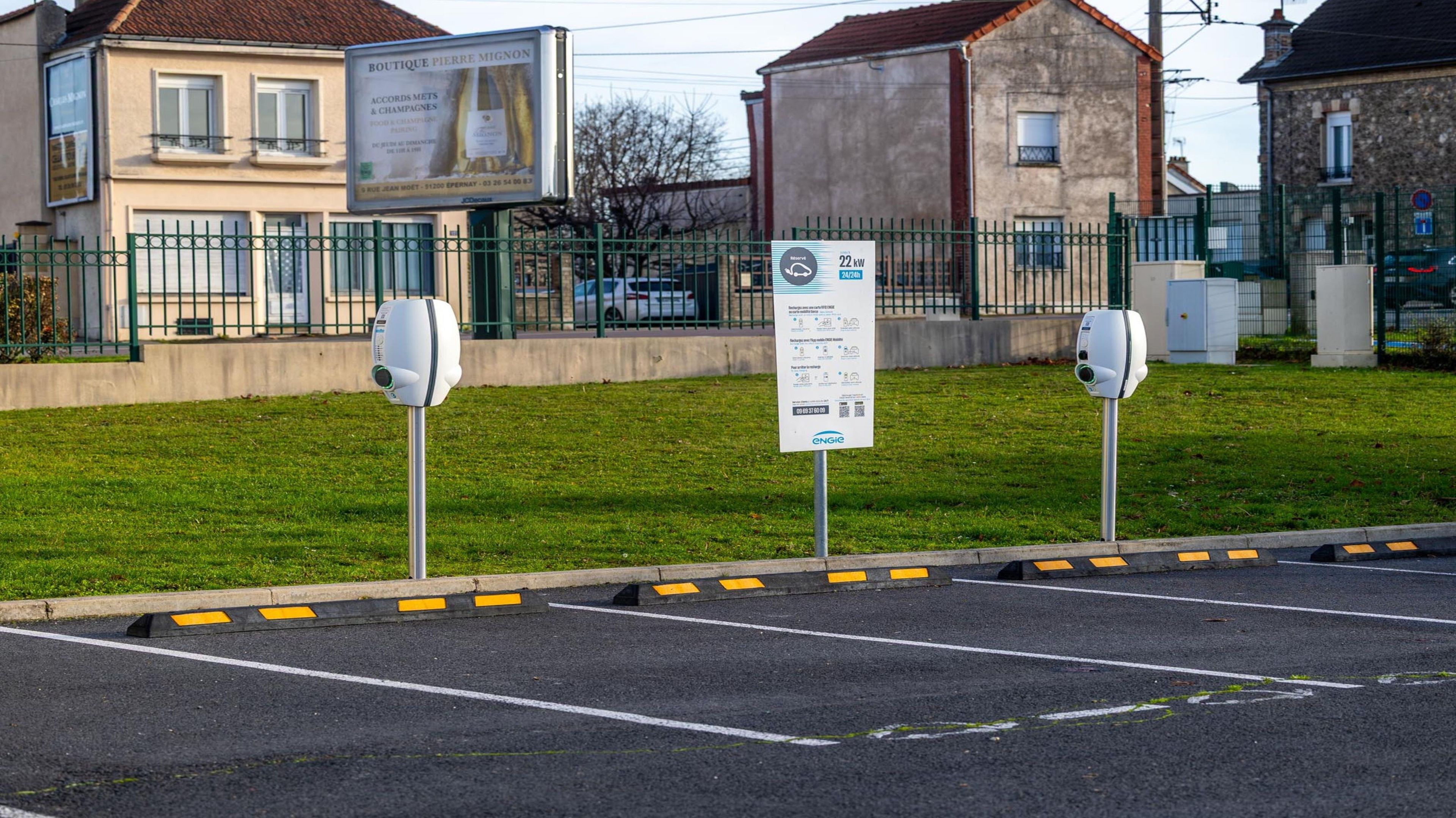 A parking area with electric vehicle charging stations, a signboard, and a building in the background.