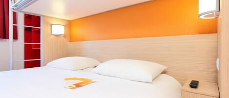 A hotel room with a bed, a nightstand, a red wardrobe, and a wall-mounted light fixture.