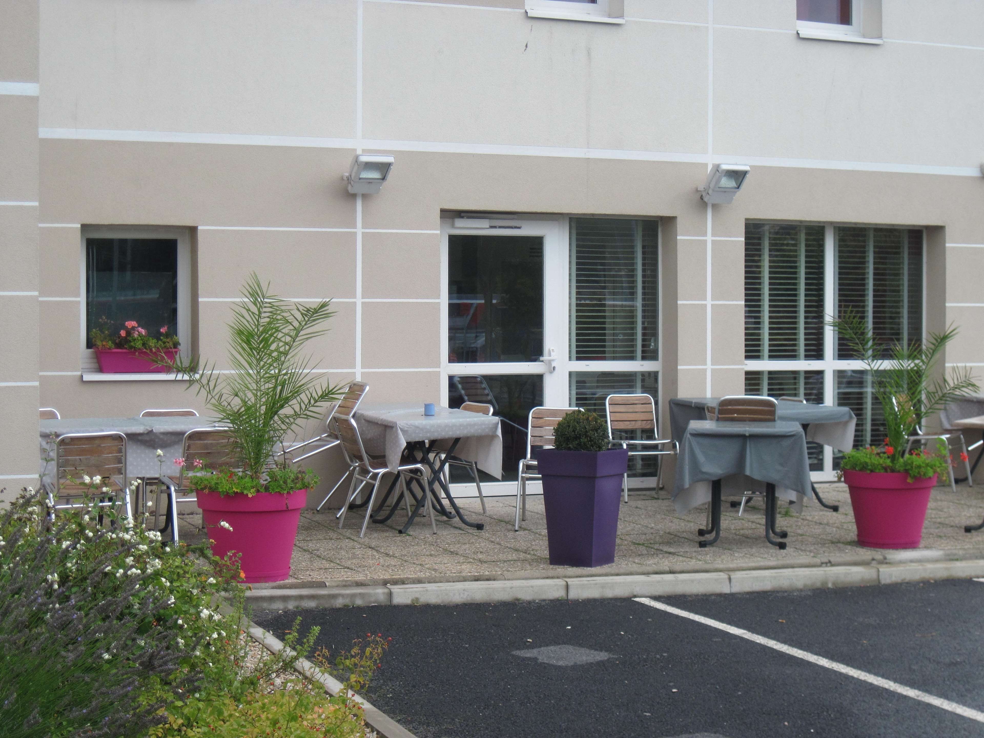 Un área de estar al aire libre con mesas y sillas, plantas en macetas y fachada de edificio con ventanas y contraventanas.