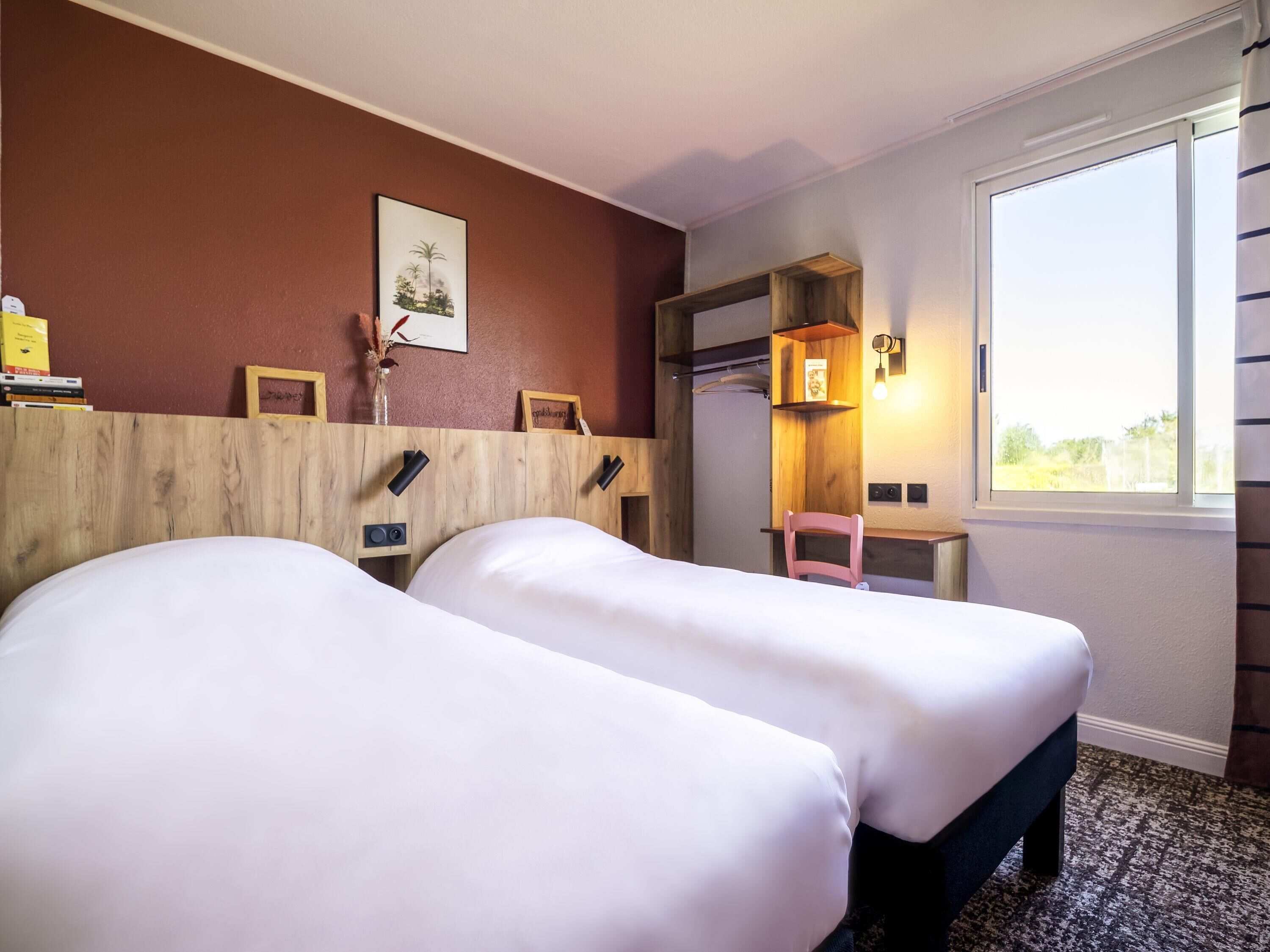 A hotel room with two beds, a wooden headboard, a small desk with a chair, a window, and a wall-mounted shelf with a picture.