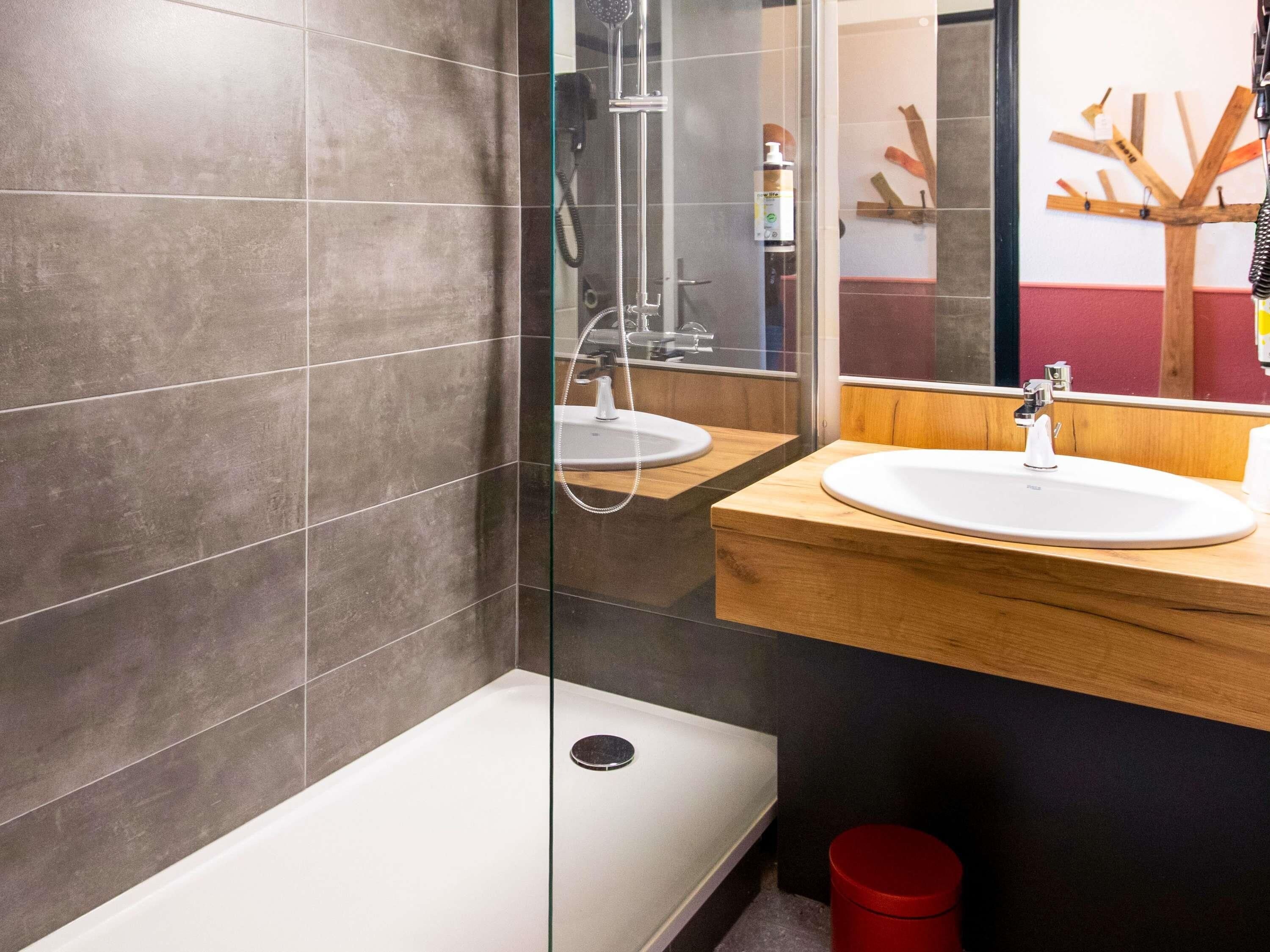 Un baño moderno con ducha de vidrio, tocador de madera con lavamanos y un bote de basura rojo.