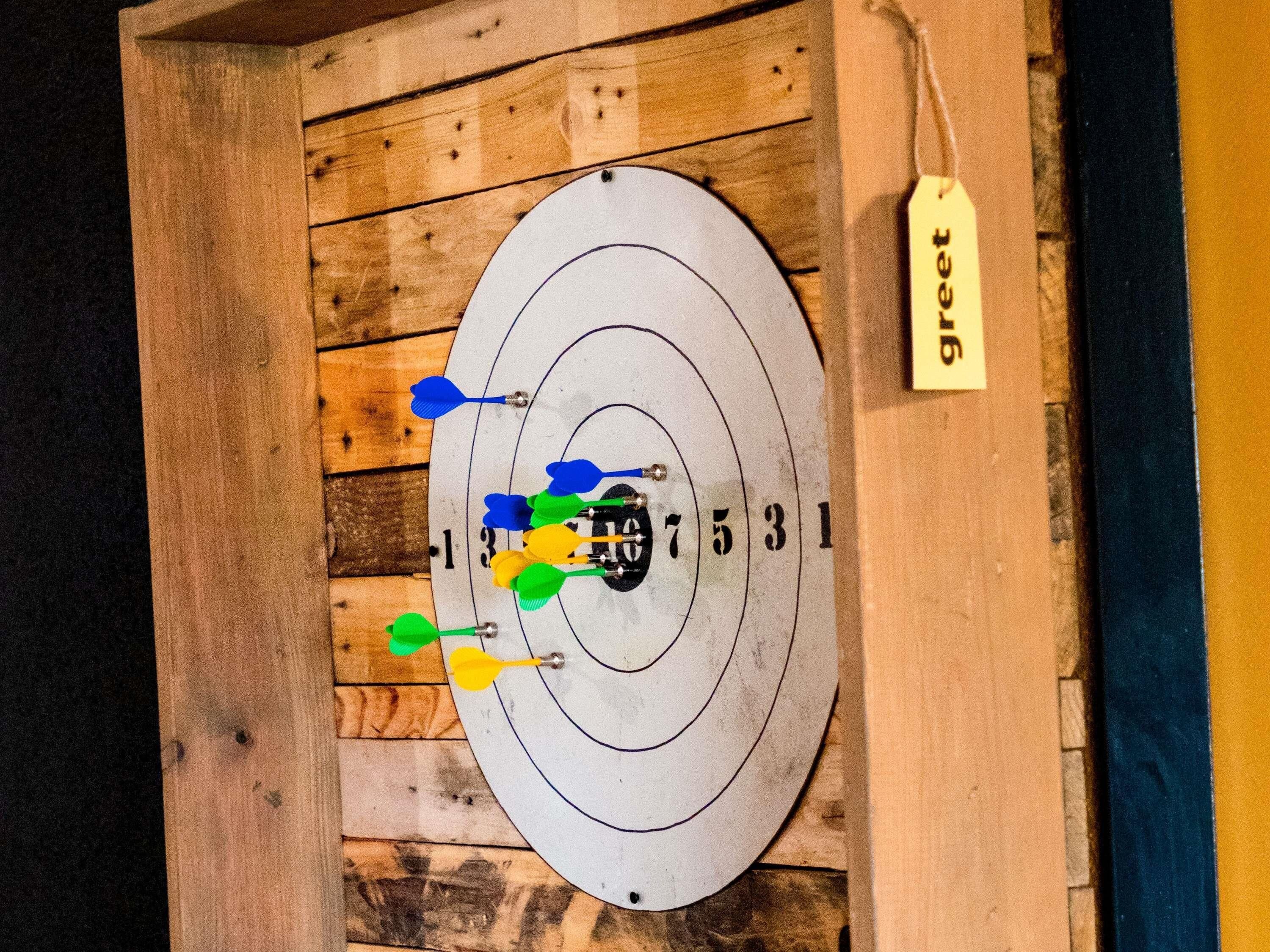 Dartbord met meerdere pijlen in verschillende kleuren, bevestigd aan een houten wand.