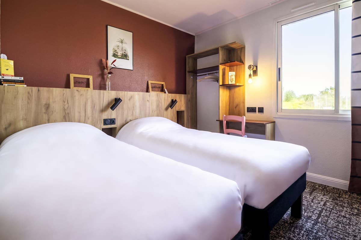 A hotel room with two beds, a wooden headboard, a small desk with a chair, a window, and a wall-mounted shelf with a picture.