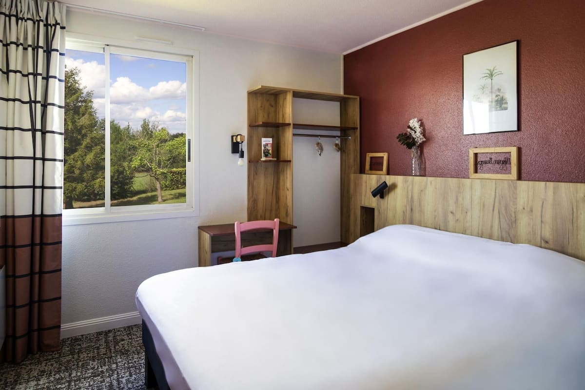 A hotel room with a bed, a desk, a chair, a window with a view of trees, and a wall with a framed picture and a small shelf.