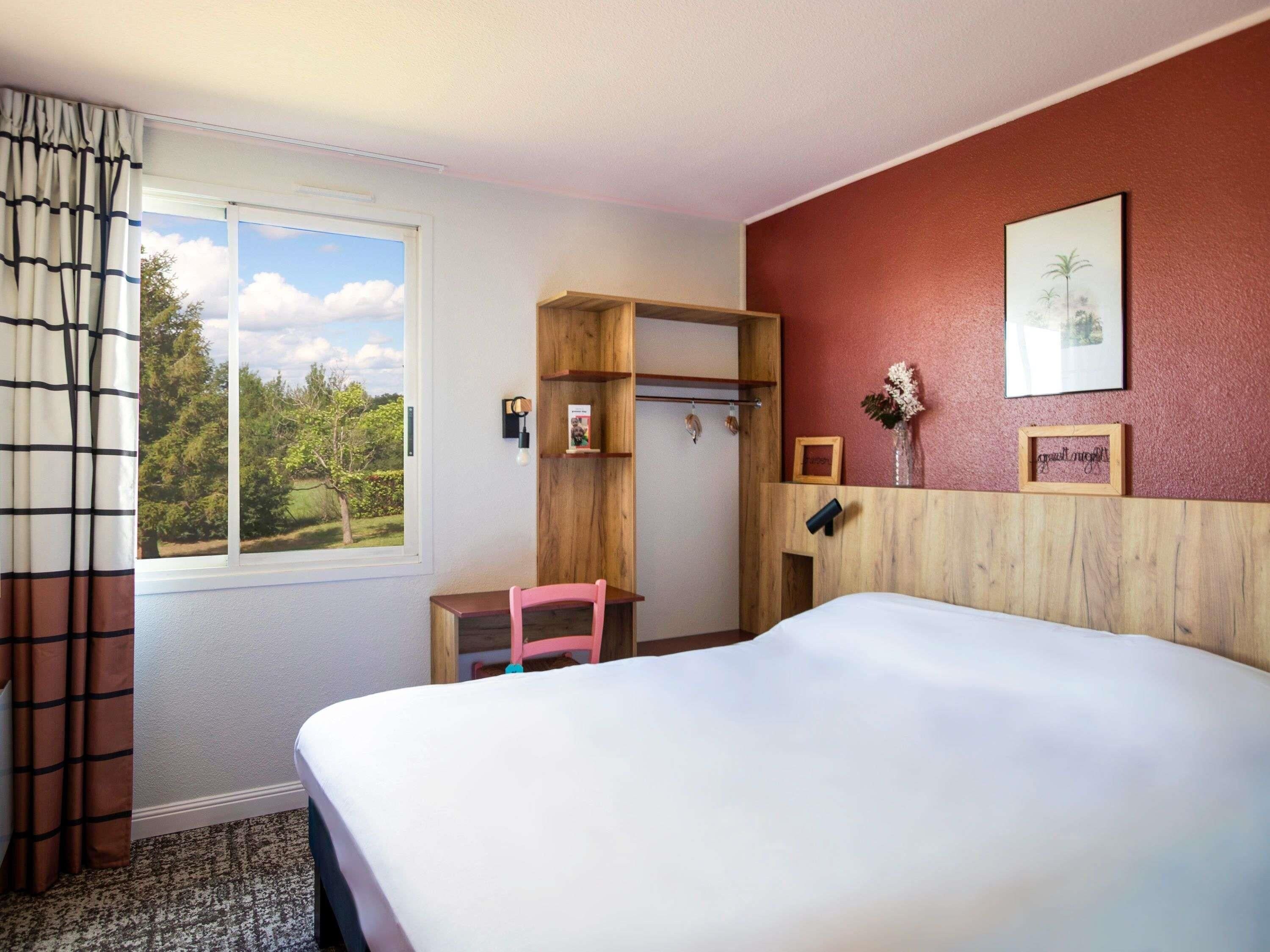 A hotel room with a bed, a desk with a chair, a window with curtains, and a view of trees.