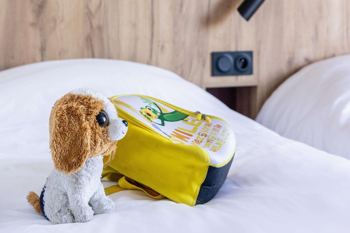 A plush toy sitting on a bed with a yellow backpack beside it.