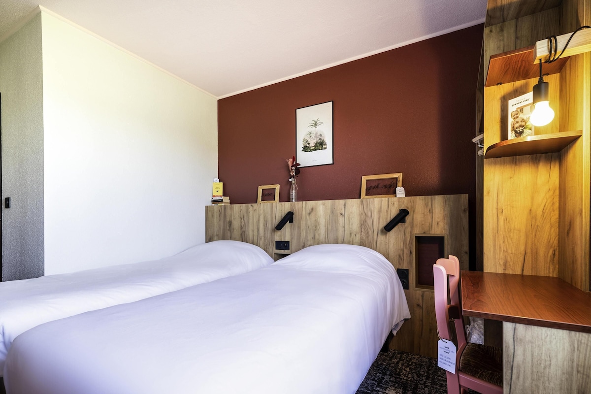 A hotel room with two single beds, a wooden desk, a chair, a small shelf with a lamp, and a framed picture on the wall.