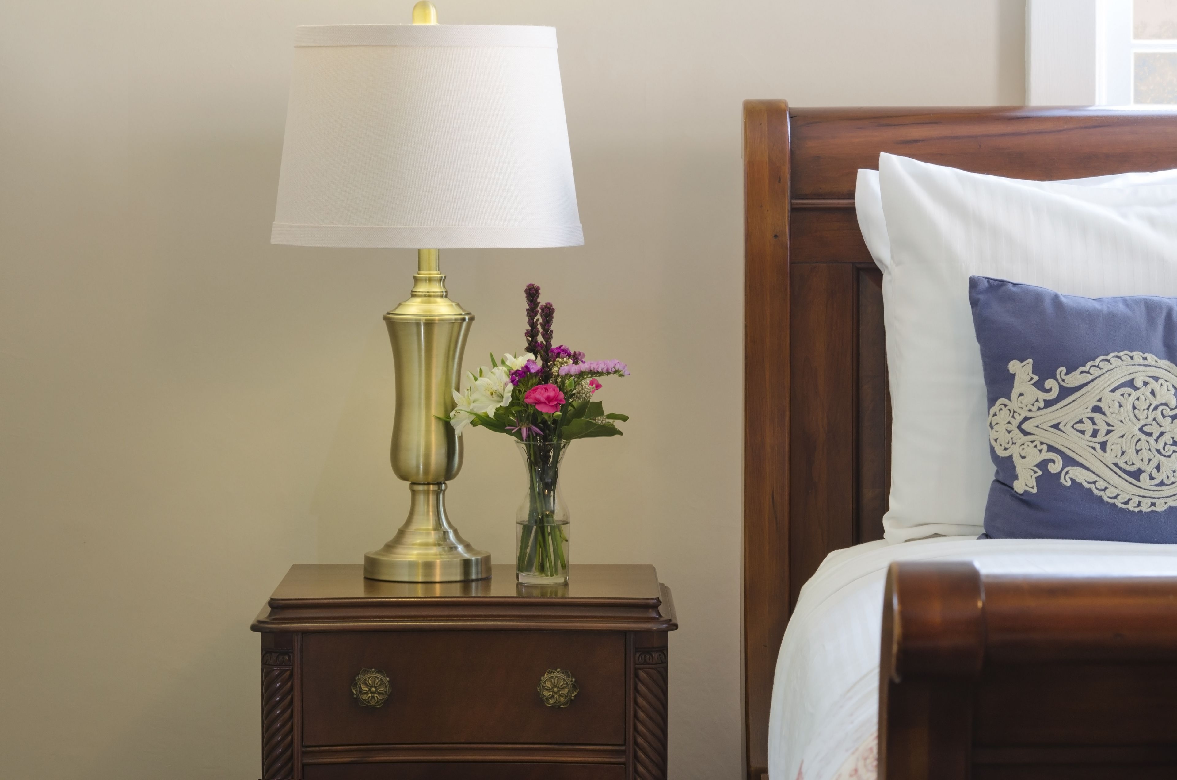 A neatly made bed with a white pillow and a decorative blue pillow, a brass table lamp, and a small vase with flowers on a wooden nightstand.