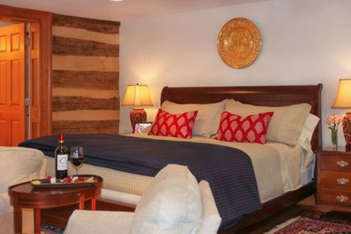 Ein Hotelzimmer mit einem hölzernen Kopfteil, einem Bett mit roten und weißen Kissen, einem Nachttisch mit einer Lampe sowie einem kleinen Tisch mit einer Weinflasche und Gläsern.