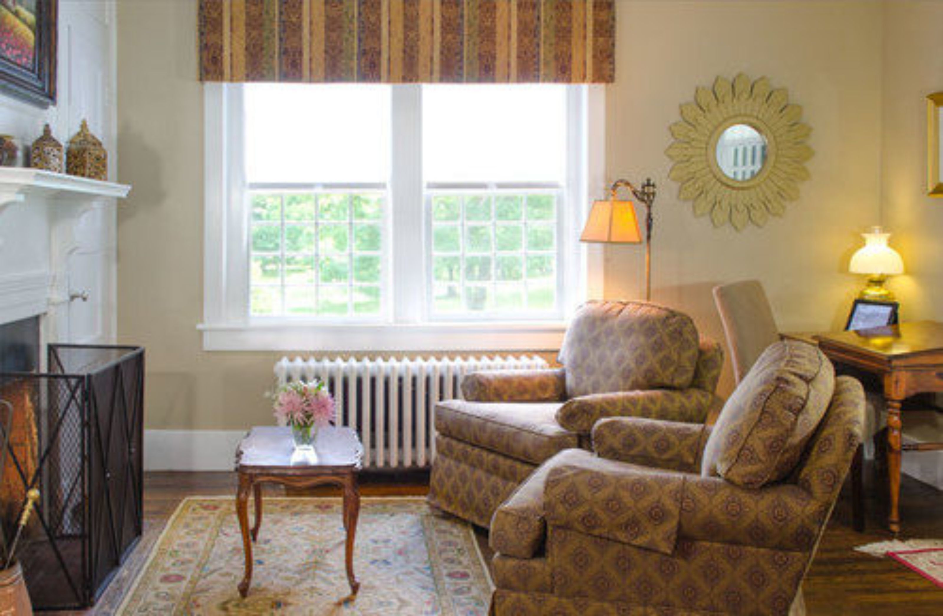A living room with a patterned armchair, a small table with a vase, a radiator, and a window with a view of greenery.