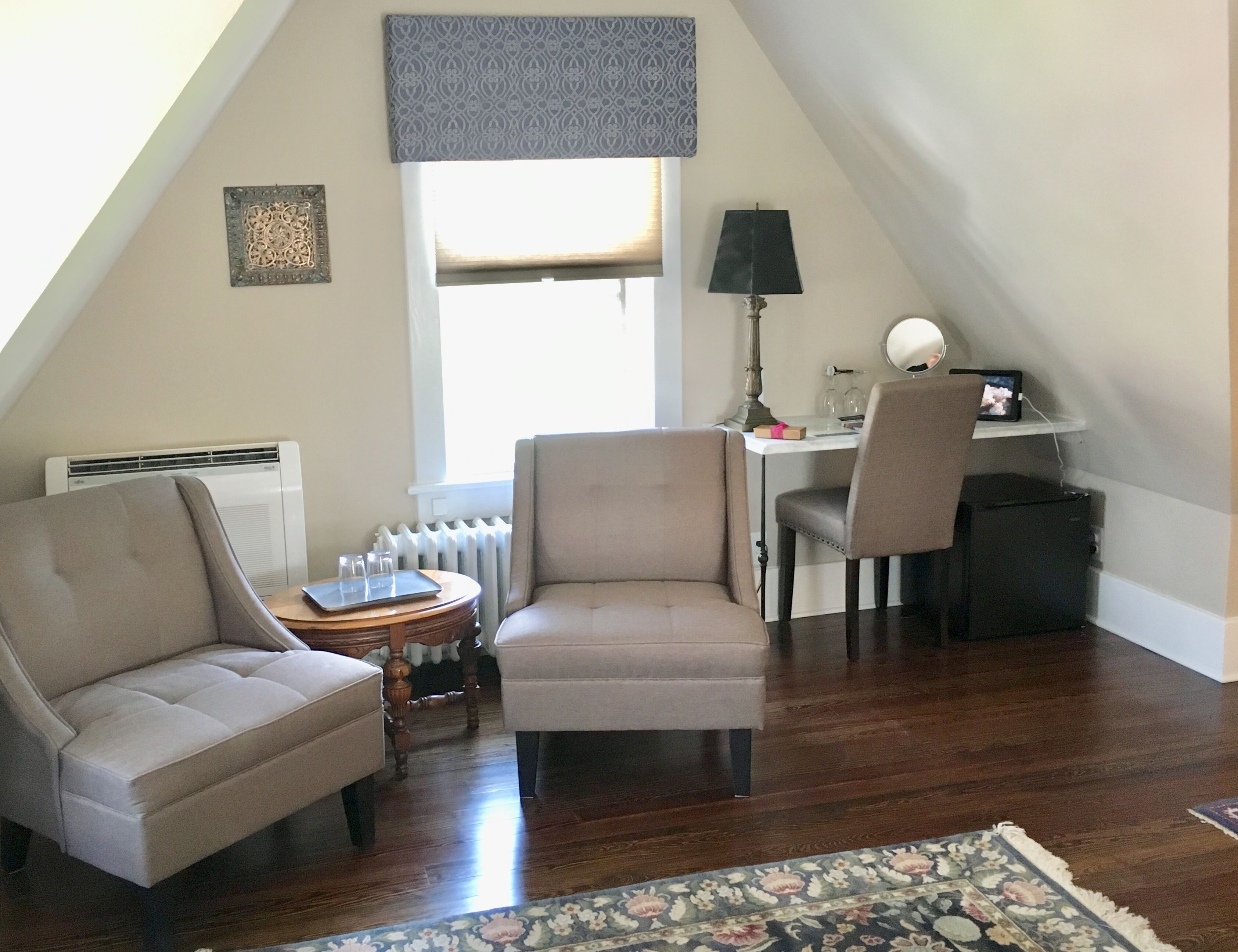 A cozy attic room with a sloped ceiling, a radiator, a desk with a lamp, two armchairs, and a small table with glasses.