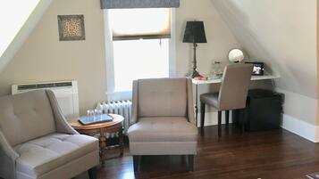 A cozy attic room with a sloped ceiling, a radiator, a desk with a lamp, two armchairs, and a small table with glasses.