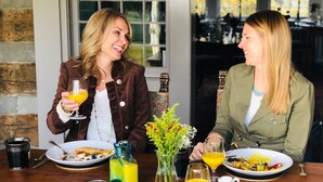 Two people sitting at a table with plates of food and drinks, a vase with flowers, and a window in the background. - Hillbrook Inn (Charles Town)