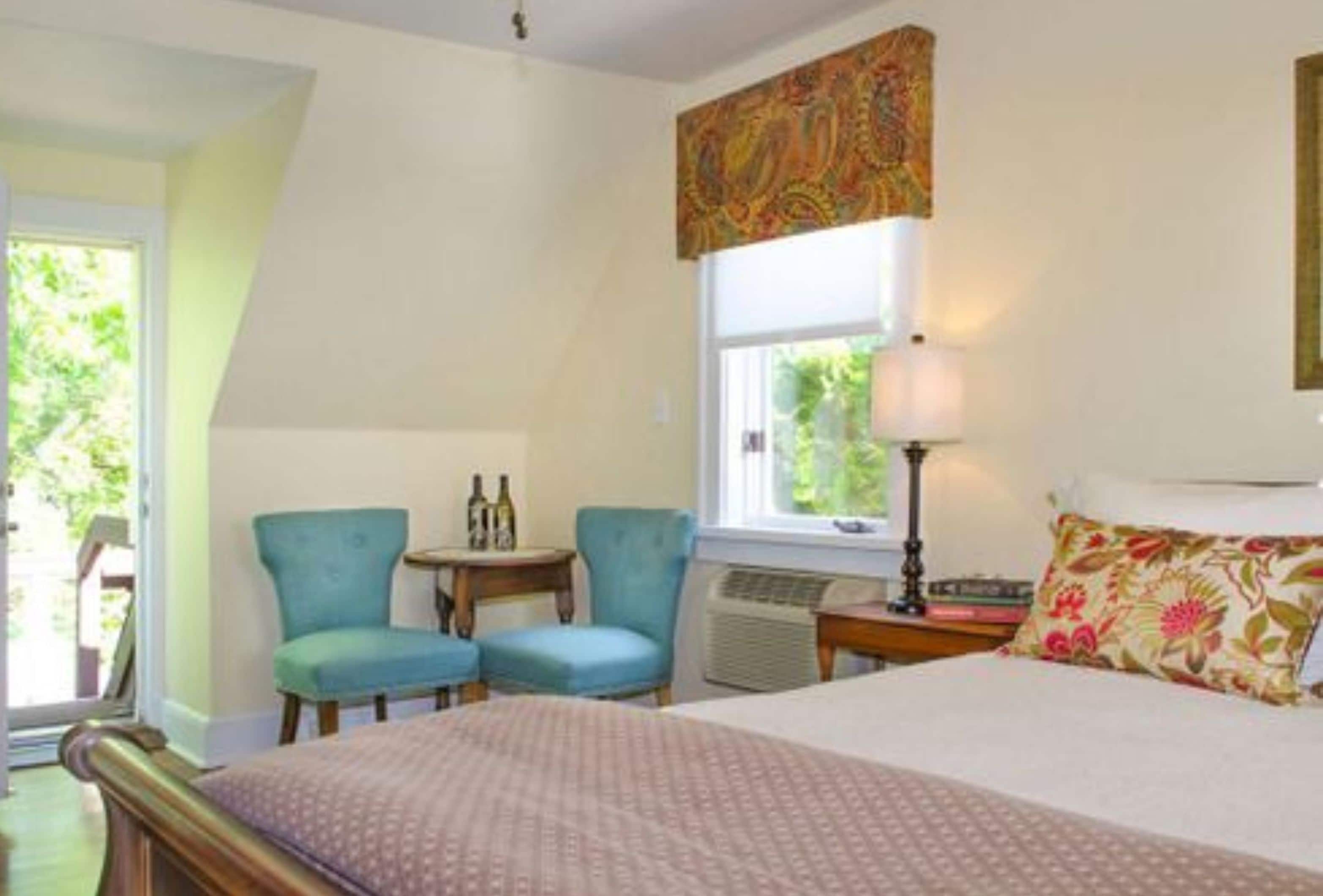 Ein Schlafzimmer mit einem Bett, zwei Stühlen, einem kleinen Tisch mit Flaschen, einem Fenster mit Vorhängen und einem Heizkörper.