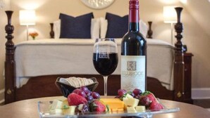 A hotel room with a bed, a bedside table with a wine bottle and glass, and a fruit platter on the table. - Hillbrook Inn (Charles Town)