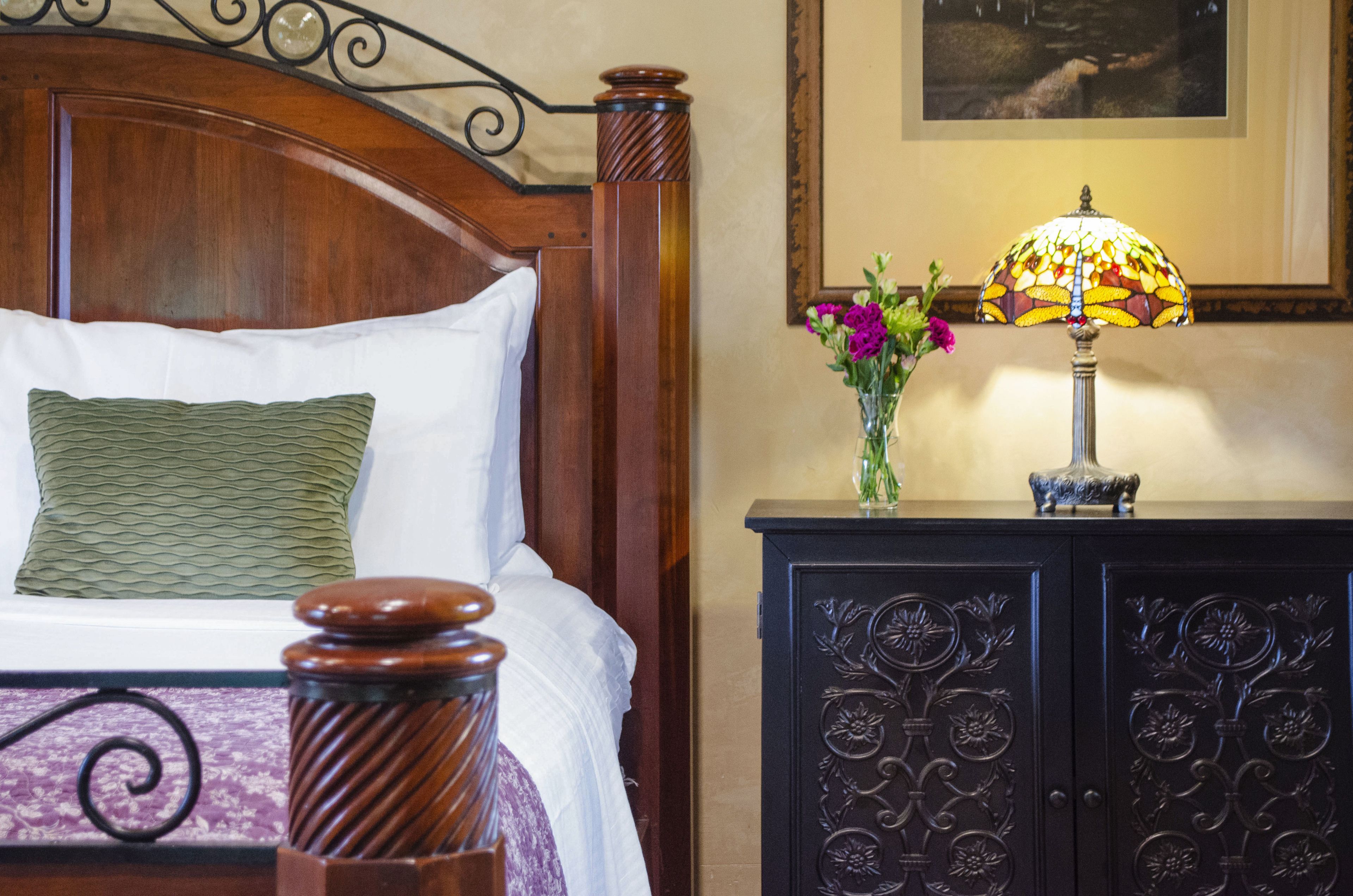 Un lit bien fait, avec une tête de lit en bois, une lampe décorative et une commode ornée de motifs floraux.