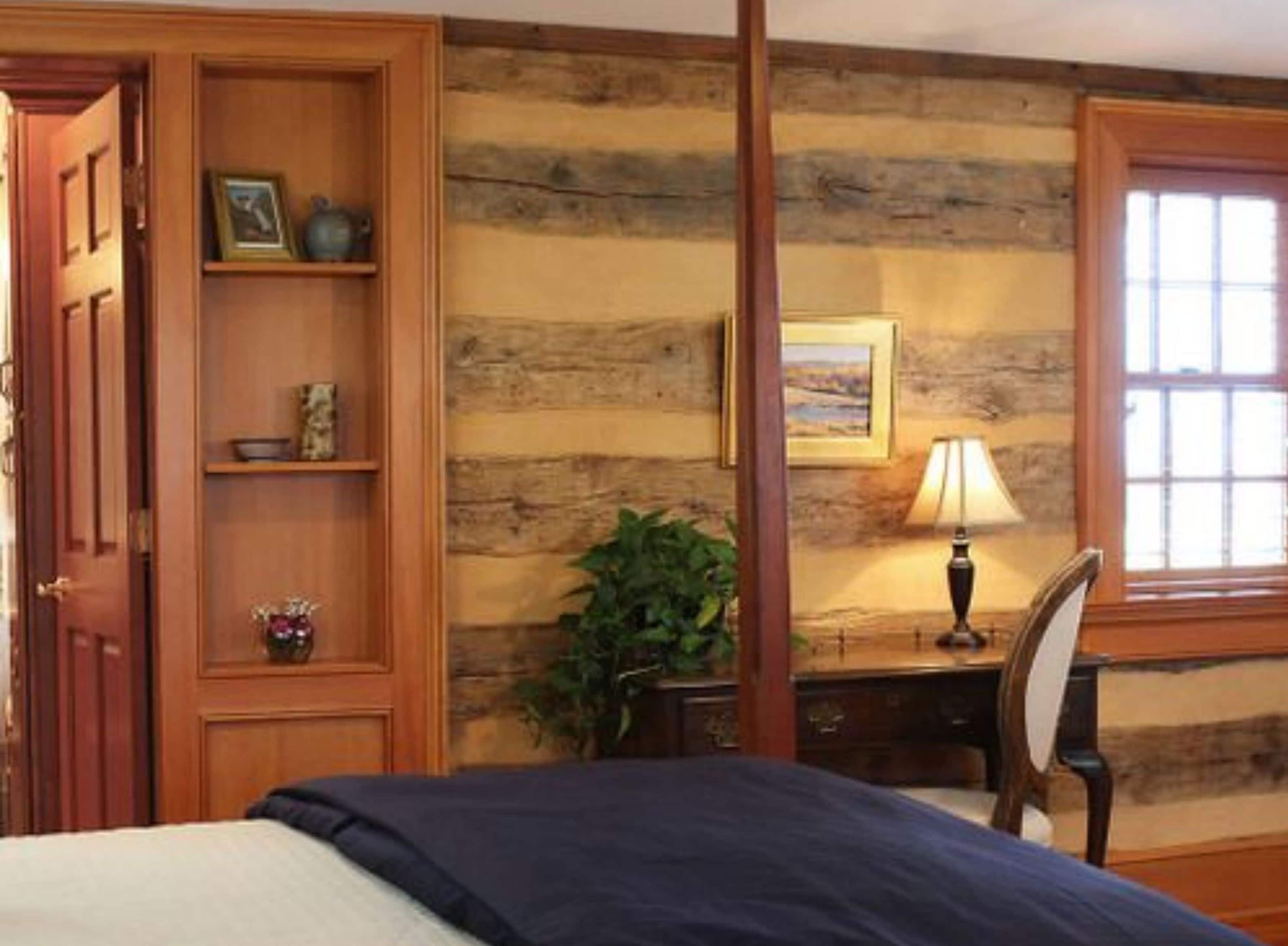 A room with a wooden wall, a bed with a blue blanket, a desk with a lamp, and a window with blinds.