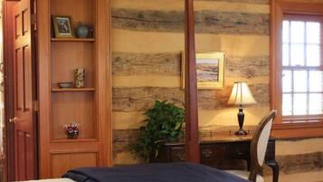 A room with a wooden wall, a bed with a blue blanket, a desk with a lamp, and a window with blinds.