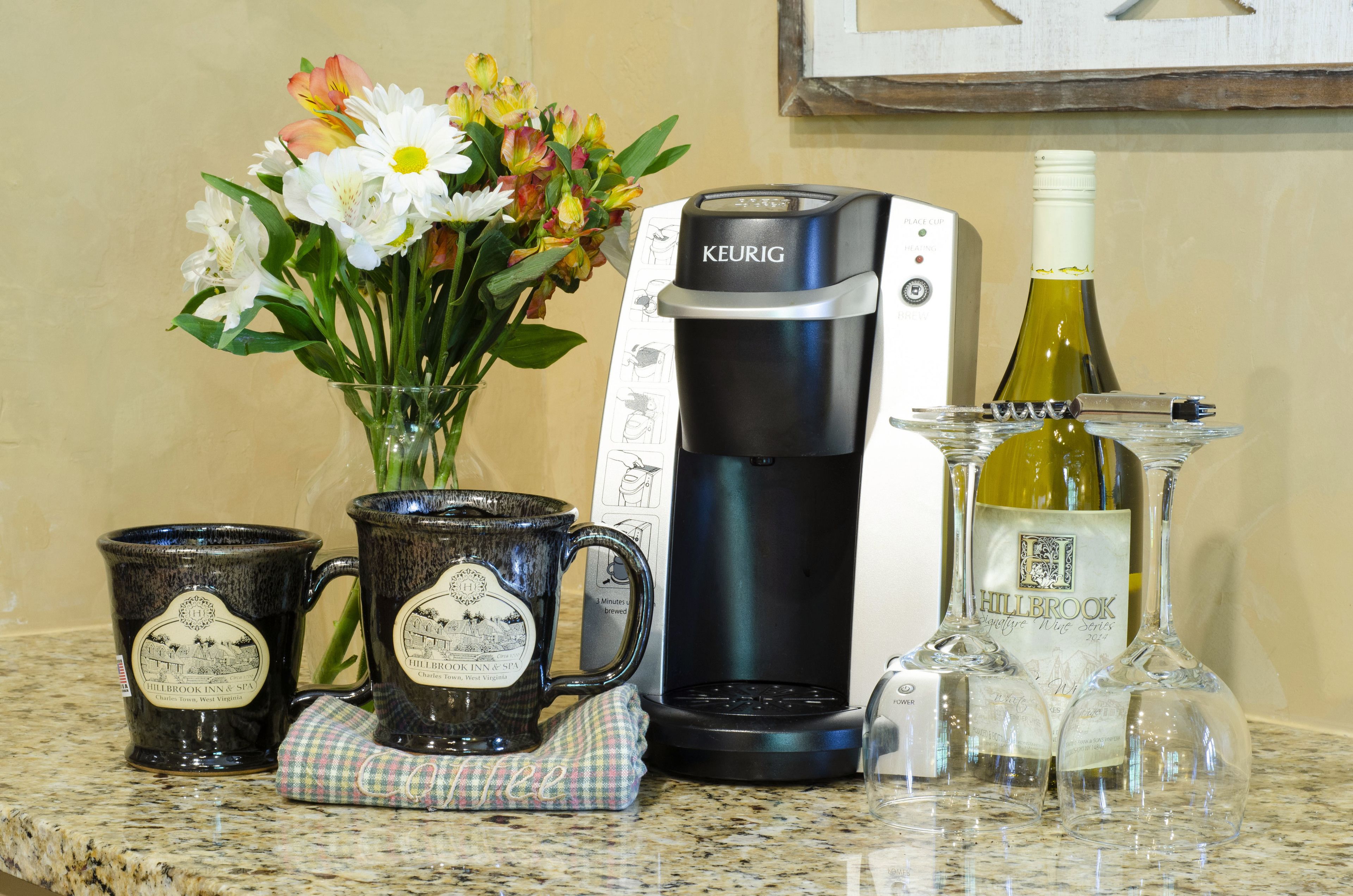 A coffee maker, two mugs, a vase with flowers, and a wine bottle on a kitchen counter.