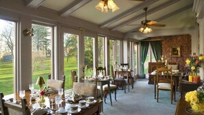 A dining room with a view of a green landscape, large windows, and a ceiling fan. - Hillbrook Inn (Charles Town)