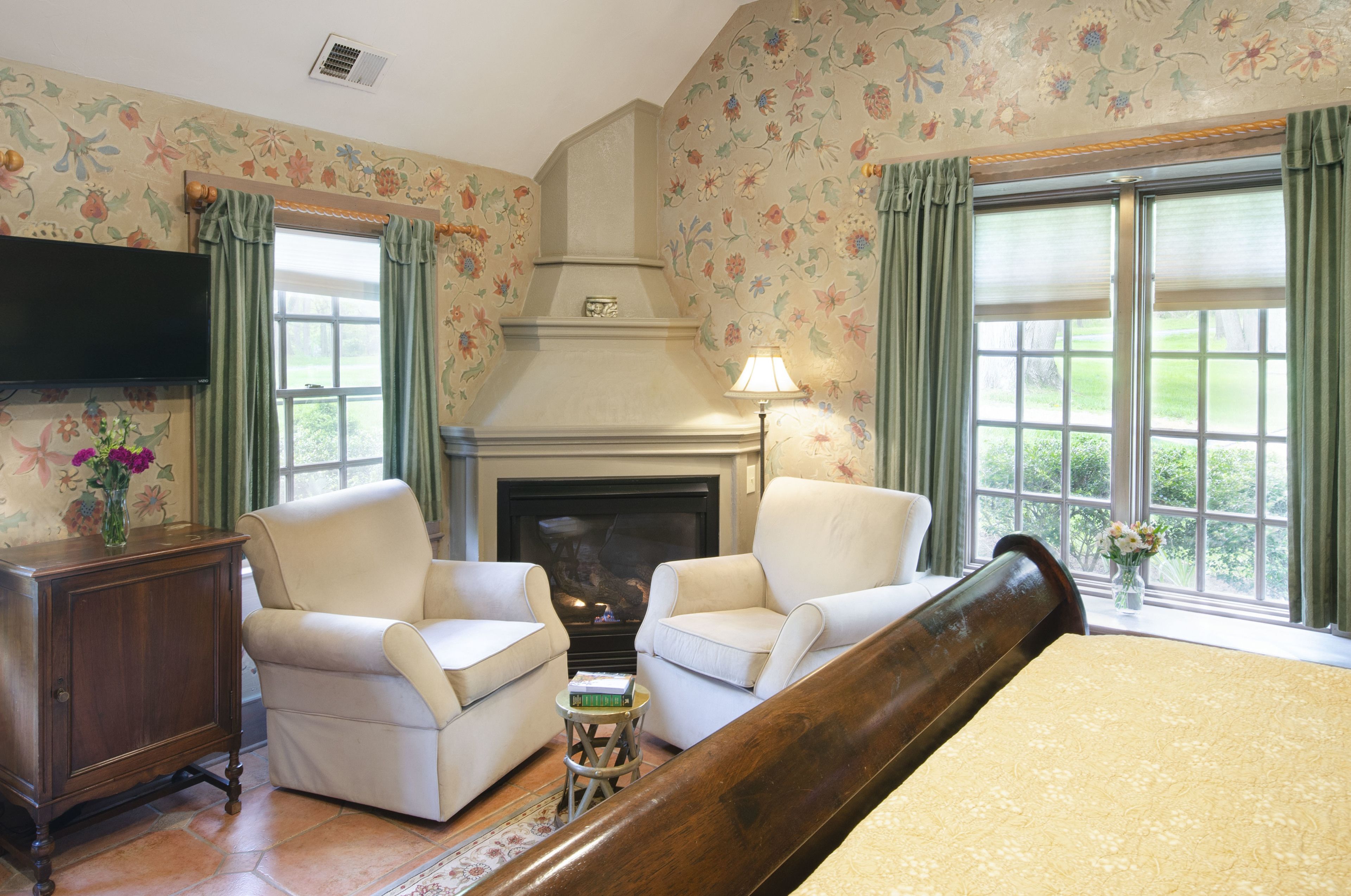 A room with two armchairs, a small table, a bed, a fireplace, and a television.