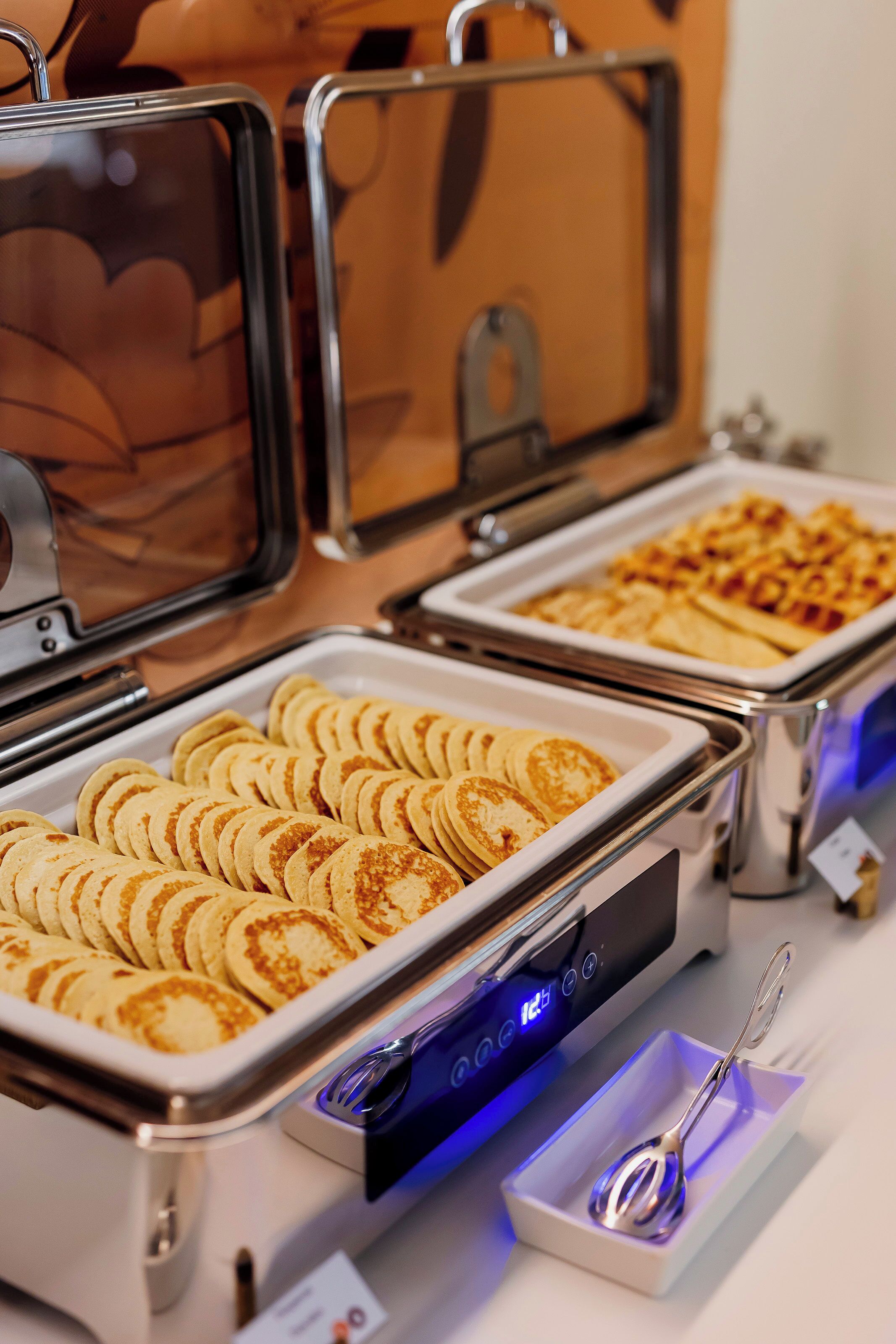 A buffet with various food items, including pancakes and waffles, served in metal trays.