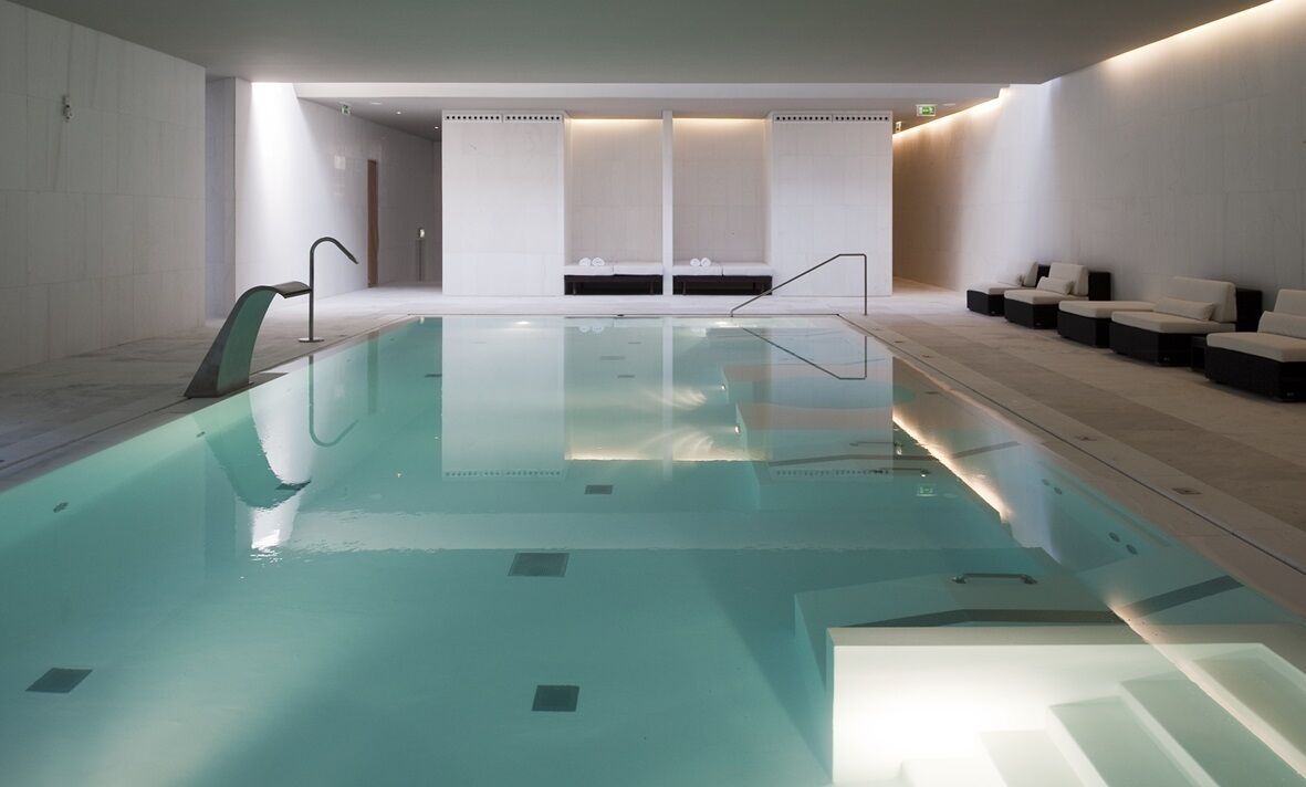 A modern indoor swimming pool with clear water, surrounded by white walls and seating areas.