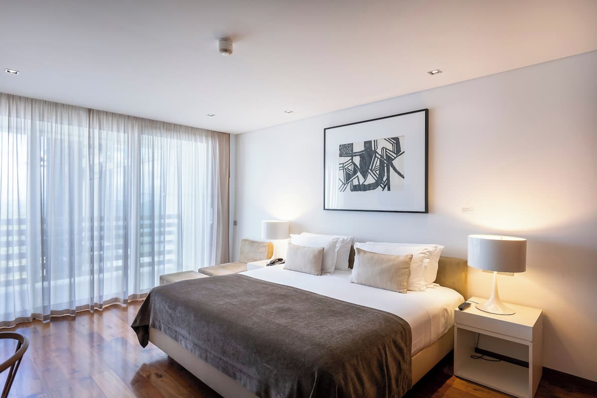 Una habitación de hotel moderna con una cama grande, dos mesitas de noche con lámparas, un ventanal con cortinas translúcidas y una obra de arte abstracta enmarcada en la pared.