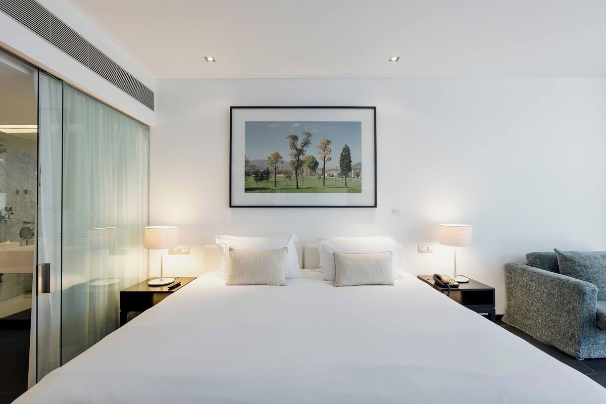 Una habitación de hotel moderna con una cama grande, dos lámparas de noche, un cuadro con un paisaje enmarcado y un sillón gris.