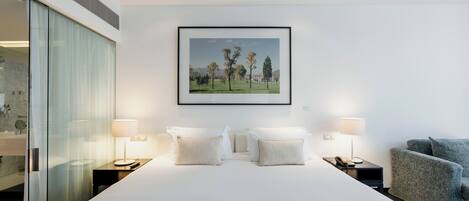 A modern hotel room with a large bed, two bedside lamps, a framed landscape painting, and a grey armchair.