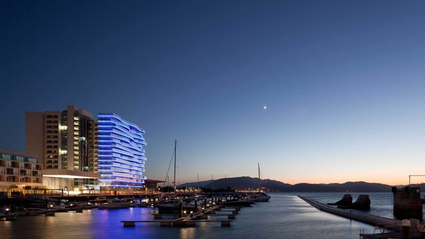 Un hotel frente al agua con diseño moderno, que presenta un edificio destacado con iluminación azul y un puerto deportivo con barcos amarrados.