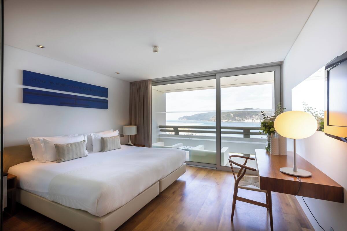 Una habitación de hotel moderna con una cama grande, un escritorio con lámpara y vistas al mar a través de un ventanal amplio.