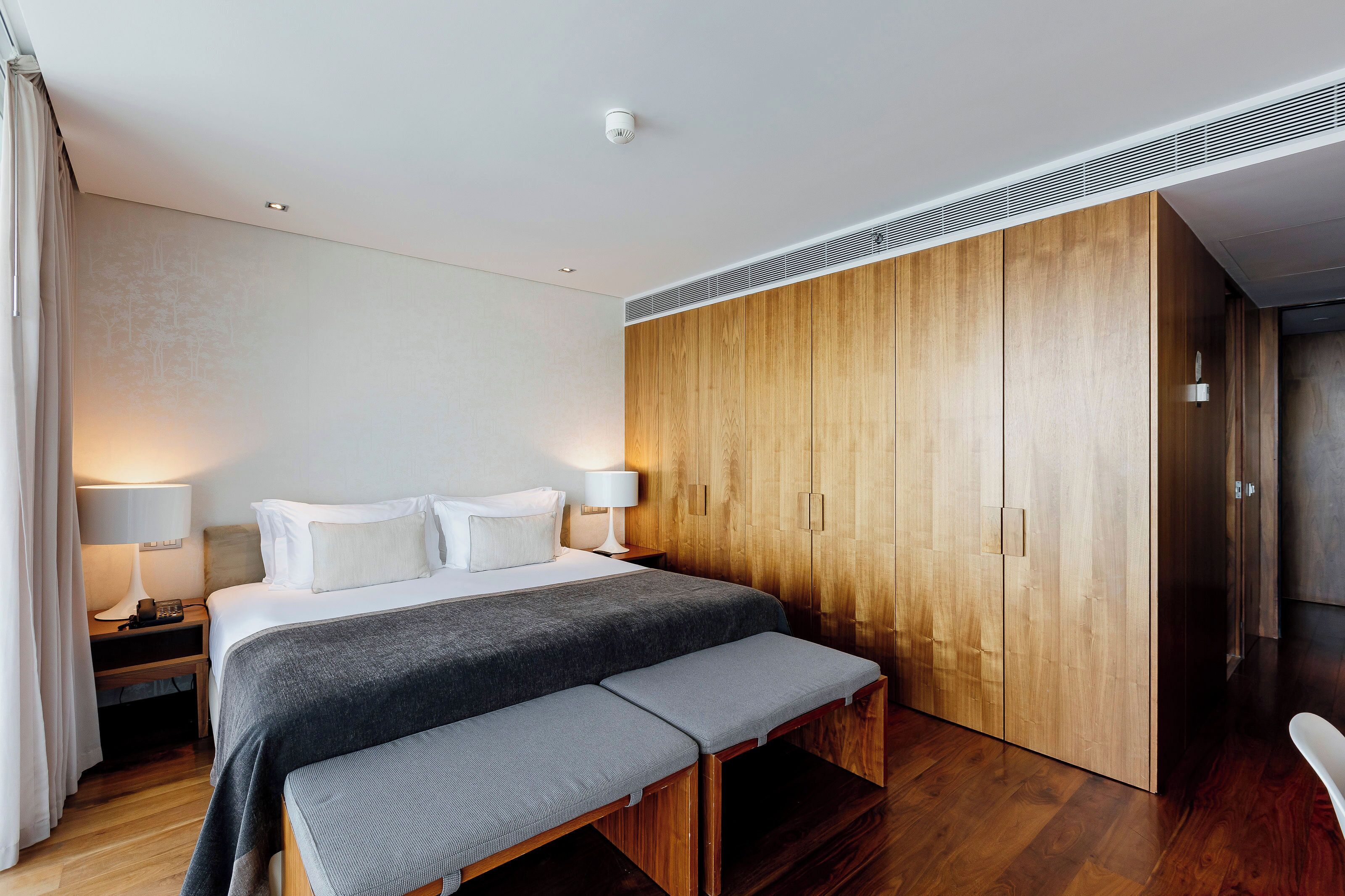 Una habitación de hotel moderna con una cama grande, lámparas de noche, un banco, un armario de madera y una rejilla en el techo.