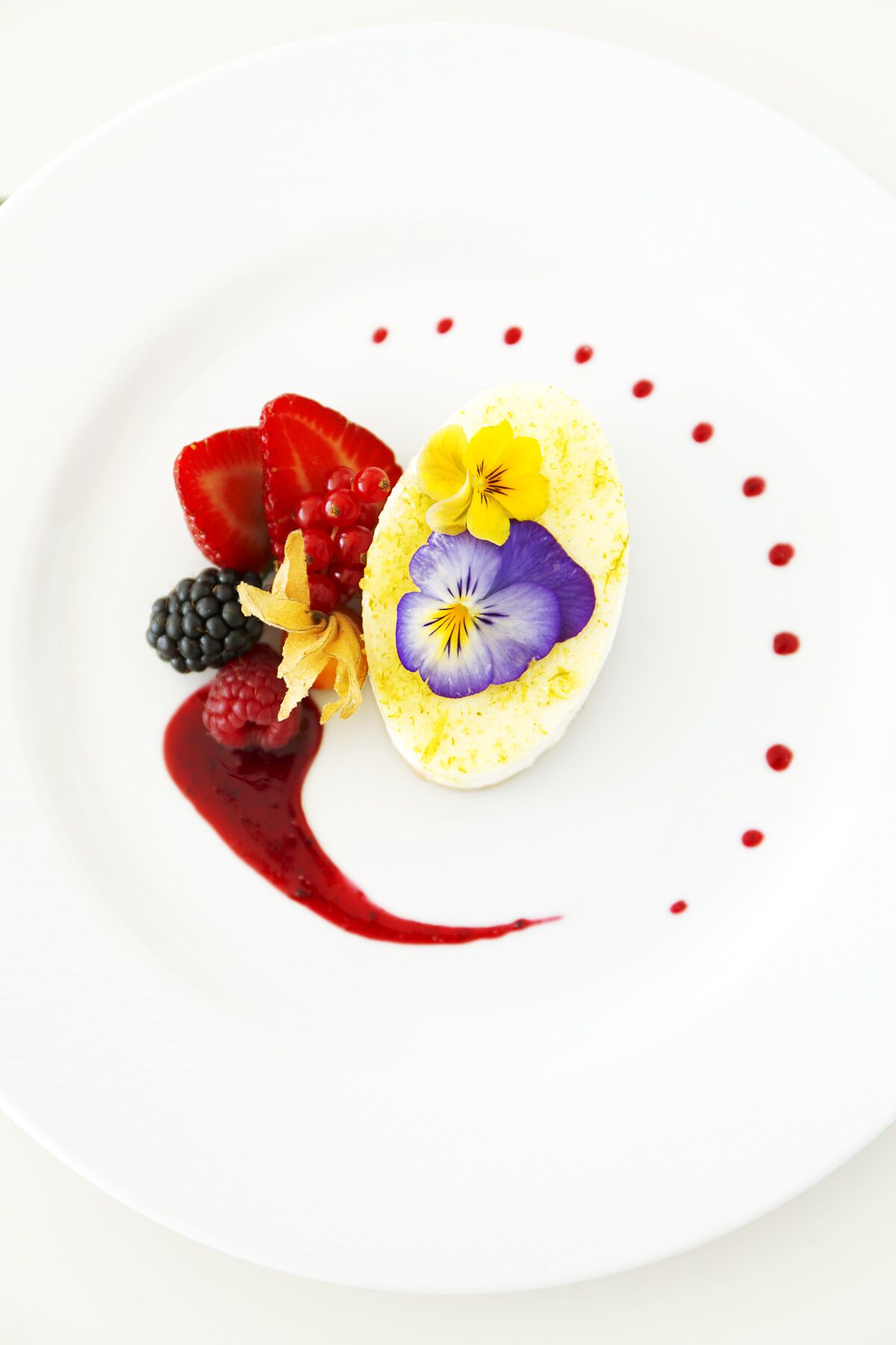 A white plate with a dessert featuring a yellow base, purple and yellow flowers, and red berries.