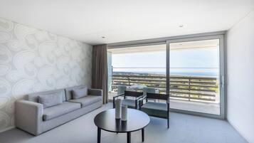 A modern hotel room with a grey sofa, a round table, and a view of a balcony with a railing.