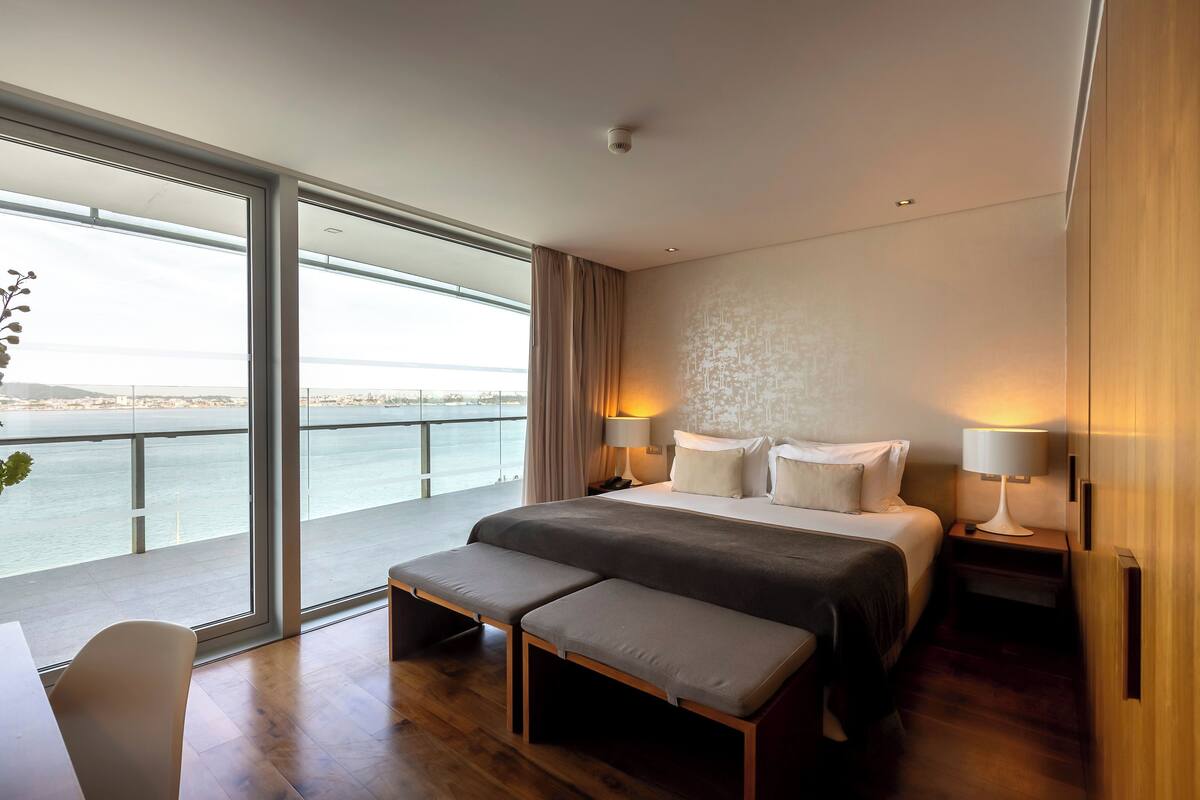 Una habitación de hotel moderna con una cama grande, lámparas de noche, un banco y vista al mar.