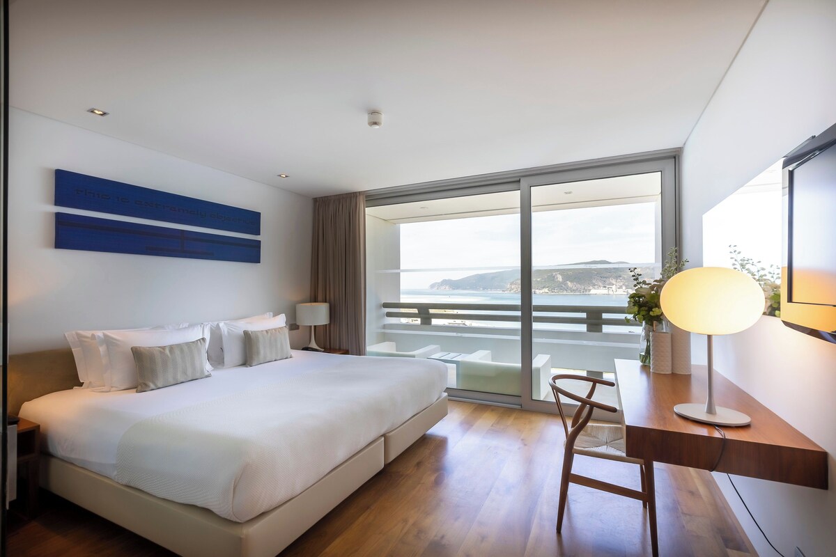 Habitació d'hotel moderna amb un llit gran, un escriptori amb una làmpada, i vista al mar a través d'una finestra ampla.