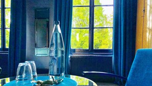 A room with a round table, a bottle, and a glass. There is a window with curtains and a view of trees outside. - Maison de Plumes (Heuchin)