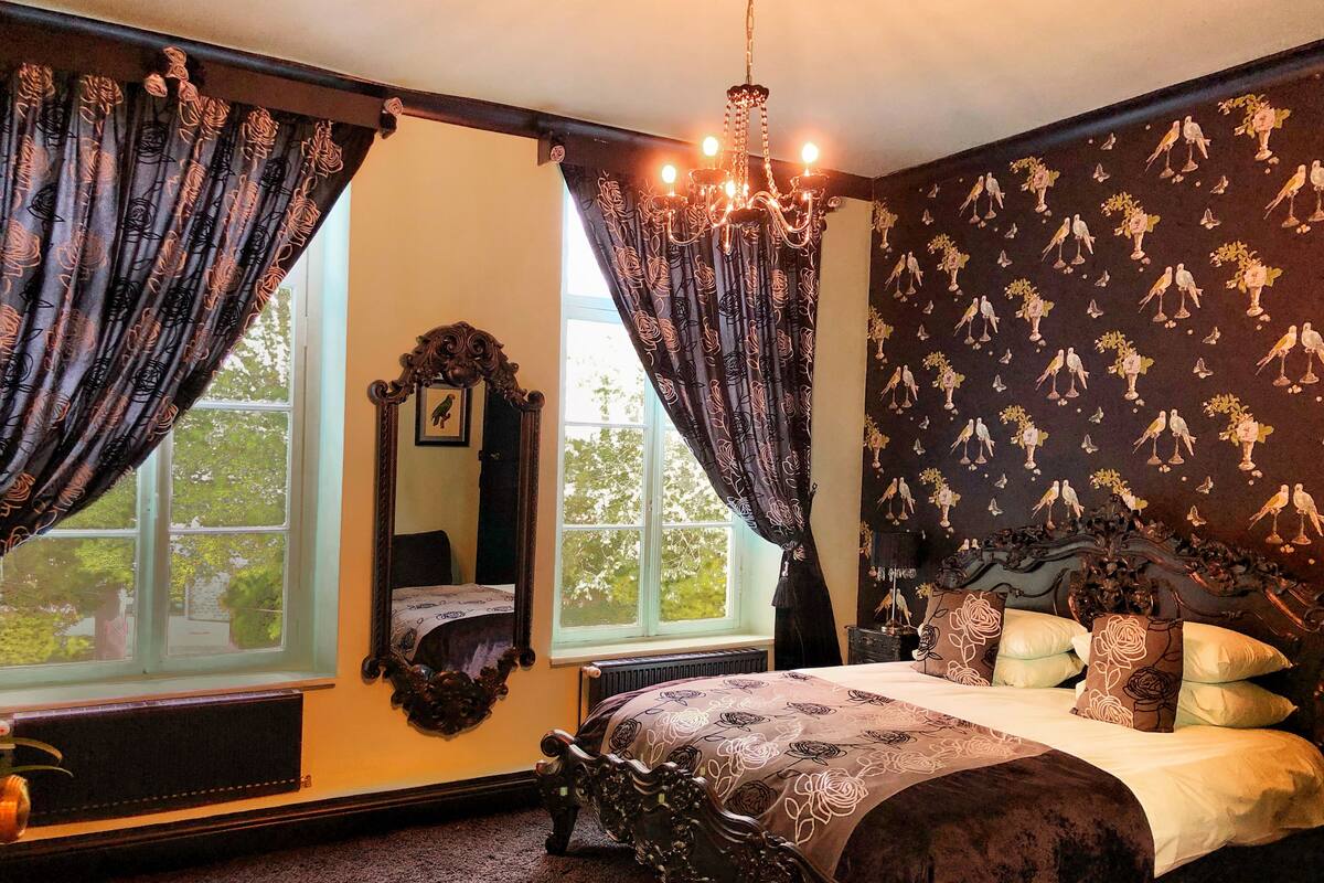 A bedroom with a large bed, ornate curtains, a chandelier, and a wall with bird patterns.