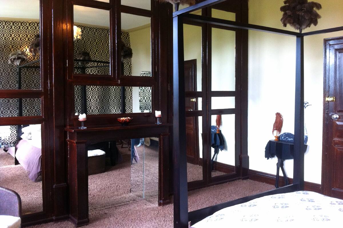 A room with a large mirror, a wooden desk, and a chair. There is a bed with a patterned bedspread and a window with a view of the outside.