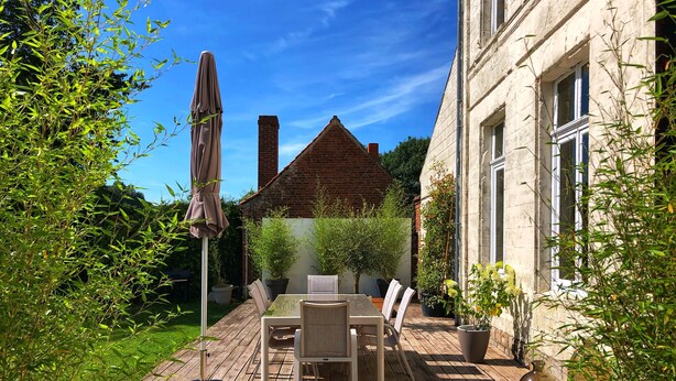 A wooden deck with a table and chairs, surrounded by greenery and a building. - Maison de Plumes (Heuchin)