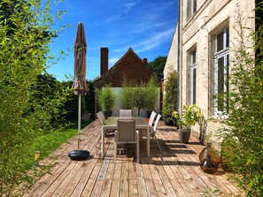 A wooden deck with a table and chairs, surrounded by greenery and a building. - Maison de Plumes (Heuchin)